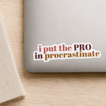 A rectangular die-cut sticker with the phrase 'i put the PRO in procastinate' in pink and brown lettering on a white background, shown applied to the lower-left corner of a silver laptop. The laptop is part of a styled flat-lay on a light wood desk, surrounded by a notebook with a gold pen, a cup of coffee, and a small green plant.