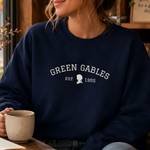 A woman wearing a navy blue crewneck sweatshirt with 'GREEN GABLES EST. 1965' printed in white on the chest, sitting in a cozy room and holding a mug.