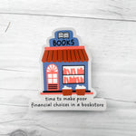 A die-cut sticker featuring a colorful illustration of a bookstore with the word 'BOOKS' above it and the phrase 'time to make poor financial choices in a bookstore' printed below, shown on a light gray wood-textured background.