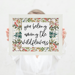 Person holding a framed print with 'you belong among the wildflowers' text and floral design.