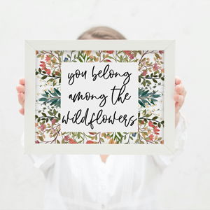 Person holding a framed print with 'you belong among the wildflowers' text and floral design.