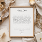 A framed typographic wall art print reading 'Apostles Creed' with the Christian creed text in black script and serif font on a white background, styled in a flat lay with a candle, ribbon, dried flowers, and a gift box.