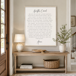 A framed wall art print featuring the Christian prayer 'Apostles Creed' in black serif text on a warm off-white background, styled in a bright, modern home interior above a wooden console table with vases and plants.