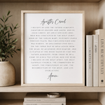 A framed typographic wall art print featuring the Apostles Creed in black script and serif text on a white background, styled on a wooden shelf beside books and a potted plant.