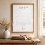 A framed personalized 'Apostles Creed' wall art print is displayed in a warm, styled home setting on a wooden console table. The print features elegant black script and serif typography on a clean white background, enclosed in a light natural wood frame.