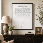 A framed typographic wall art print titled 'Apostles Creed' hangs on a beige wall above a dark wooden console table. The print features black text on a white background with a thin black frame.