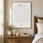 A framed vertical wall art print featuring the Apostle Creed in black serif text on a warm off-white background, styled in a bedroom setting above a wooden nightstand with a vase, candle, and bed.