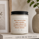 A white glass candle jar with a black lid sits on a shelf beside books, a framed botanical print, and a beige vase with green leaves. The candle label reads, 'Our love is like this candle.
