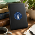 A black rectangular device or case standing upright on a wooden desk, featuring a circular blue and white sticker that reads 'THAT'S DEBATABLE' with a podium icon. The scene includes stacked books, a plant, a cup of coffee, and a notebook in a styled indoor setting.