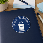 A navy blue binder or folder on a wooden desk, featuring a circular white graphic with a podium and the words 'THAT'S DEBATEABLE.' Surrounding props include a small plant, stacked papers, a pen, sticky notes, and another pen.