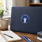 A navy blue laptop sits on a wooden desk in a bright room, featuring a circular blue and white sticker that reads 'THAT'S DEBATABLE' with a podium icon. An open notebook, pens, and a coffee mug are arranged around it.