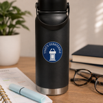 A matte black insulated water bottle with a carry loop and stainless steel base, standing on a wooden desk. The bottle has a blue circular sticker that reads 'THAT'S DEBATABLE' with a podium icon.