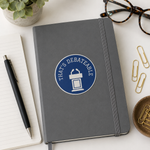 A dark gray hardcover notebook with an elastic closure, featuring a blue circular sticker that reads 'THAT'S DEBATABLE' with a podium graphic. The notebook is styled in a flat-lay arrangement on a white desk alongside a black pen, an open lined notebook, tortoiseshell eyeglasses, paper clips, and a small potted succulent.