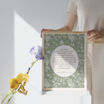 Person holding a framed print with floral design and text, next to a vase with flowers on a white background