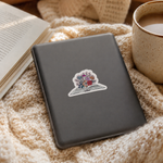 A dark gray laptop resting on a cozy beige knitted blanket, with an open book and a cup of coffee nearby. The laptop has a small die-cut sticker on the lid featuring a colorful open book with flowers.
