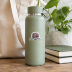 A matte sage green insulated water bottle with a loop handle and a small sticker of an open book with colorful flowers, styled on a wooden table next to a canvas tote bag, a potted plant, and stacked books.