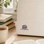 A small die-cut sticker featuring a colorful bouquet of flowers emerging from an open book, placed on the corner of a cream-colored laptop on a wooden desk. The scene is styled with a mug of tea, stacked books, and an open book in a bright, natural setting.