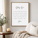 A framed wall art print featuring the Bible verse 'Glorify Be' in black script and serif text on a white background, styled in a warm living room setting next to a sofa and a side table with a candle and plant.