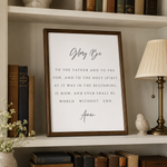A framed typographic wall art print with the Holy Trinity quote in black script and serif text on a white background, displayed on a bookshelf beside a lamp, books, and a small bird figurine.