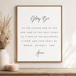 A framed typographic wall art print featuring the Bible verse 'Glory Be' in black script and serif text on a white background, hanging on a beige wall above a wooden surface with a candle and dried grass in a vase.