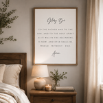 A framed wall art print featuring the Bible verse 'Glorify Be' in elegant black typography on a white background, styled in a warm, neutral-toned bedroom setting above a wooden nightstand.