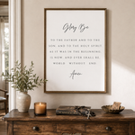 A framed wall art print featuring the Bible verse 'Glorify Be to the father and to the son, and to the Holy Spirit, as it was in the beginning, is now, and ever shall be, world without end. Amen' from Revelation, styled on a wooden console table with a candle, prayer beads, a book, and a bowl in a warm, neutral room.