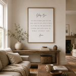 A framed wall art print featuring the Bible verse 'Glorify Be' with elegant black script and serif text on a white background, styled in a warm, modern living room setting above a wooden console table.