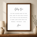 A framed typographic wall art print featuring the Bible verse 'Glory Be' in black script and serif text on a white background, shown hanging on a beige wall above a wooden console with a lamp and vase.