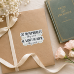 A kraft paper gift box tied with a cream ribbon, featuring a die-cut sticker that reads 'GO TO PEMBERLEY, THEY SAID HE WON'T BE HOME, THEY SAID' in black lettering with small green floral accents. The box is styled on a light beige surface with white baby's breath flowers, pink roses, and a vintage green book titled 'Pride and Prejudice'.