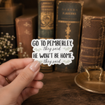 A hand holds a die-cut sticker that reads 'GO TO PEMBERLEY, they said HE WON'T BE HOME, they said' with small floral accents. The background shows a warm, blurred scene of vintage books on a wooden shelf.