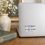 A white die-cut sticker with a black typewriter font quote reading 'GO TO PEMBERLEY, they said HE WON'T BE HOME, they said' is shown on the back of a silver laptop. The laptop sits on a wooden table next to a vintage book and a ceramic vase of white flowers.