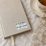 A beige linen-covered notebook with a ribbon bookmark lies on a light fabric surface beside a teacup and saucer and dried baby's breath flowers. A die-cut sticker on the notebook reads, 'GO TO PEMBERLEY, they said HE WON'T BE HOME, they said.