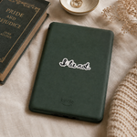A dark green Kindle tablet case with a white 'I lie not.' sticker and a gray Kindle logo, styled on a beige fabric surface next to a book, a bowl, and dried flowers.