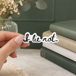 A hand with pale pink manicured nails holds a die-cut vinyl sticker that reads 'I lie not.' in black cursive script with a white outline.