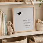 A framed nursery art print with a black heart and the text 'you have filled my heart with greater joy psalm 1:7' displayed on a shelf next to a plush bunny, books, and woven baskets.