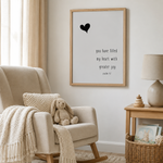 A framed wall art print with a black heart and the text 'you have filled my heart with greater joy psalm 7:1' hanging in a softly styled nursery or living room setting with a beige armchair, throw blanket, bunny toy, side table, and lamp.