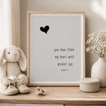 A framed typographic art print with a black heart and the quote 'you have filled my heart with greater joy psalm 1:7' displayed on a wooden surface with a plush bunny, baby shoes, and a vase of baby's breath.