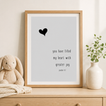 A framed typographic wall art print with a black heart and the quote 'you have filled my heart with greater joy psalm 7:1' on a light gray background, displayed in a light wood frame on a wooden surface beside a plush bunny and a small vase with greenery.