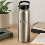 A stainless steel water bottle with a black lid and handle, featuring the printed phrase 'I lie not.' in black script with a white outline, set on a wooden desk surrounded by books and a potted plant.