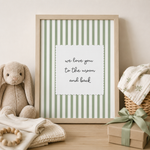 A framed nursery print with green and white vertical stripes and a central white card reading 'we love you to the moon and back,' styled on a wooden surface with a beige bunny toy, folded blankets, and a gift box.
