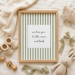 A framed baby shower print with green and white vertical stripes and the text 'we love you to the moon and back' in the center, styled in a flat lay with baby accessories including knit booties, a wooden rattle, a plush bunny, and dried flowers on a light fabric background.