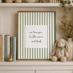 A framed vertical art print with green and white stripes and the text 'we love you to the moon and back' displayed on a shelf next to books, a bunny plush, and dried flowers.