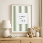 A framed vertical art print with sage green and white stripes and a central white card reading 'we love you to the moon and back,' displayed on a light wood dresser above a lamp, books, a teddy bear, and a small vase of dried flowers.