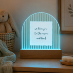 An illuminated acrylic night light with a wooden base sits on a wooden table in a softly styled room. The light has vertical blue and white stripes with a central white square that reads 'we love you to the moon and back' in dark script.