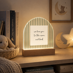 A decorative illuminated tabletop lamp shaped like a crescent moon with pale green vertical stripes and a central white card that reads 'we love you to the moon and back.' It sits on a wooden shelf in a cozy room setting with books, a plush toy, and a moon-and-star night light nearby.