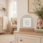 A nursery decor scene with a wooden block sign featuring a green and white striped arch and a white center card that reads, 'we love you to the moon and back.' The sign sits on a light wood dresser in a softly styled nursery with a crib, chair, stuffed bunny, and framed wall art in the background.