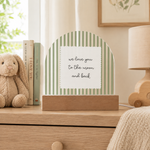 A decorative arched tabletop sign with pale green vertical stripes and a white center panel reading 'we love you to the moon and back,' displayed on a wooden dresser in a softly styled room with a bunny toy, books, a lamp, and framed wall art.
