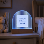 A glowing LED night light shaped like a semi-circular arch with blue and white vertical stripes, featuring a white insert with the handwritten phrase 'we love you to the moon and back'. It sits on a wooden bedside table next to a teddy bear and books in a cozy bedroom setting.