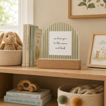 A wooden arch-shaped acrylic plaque with green and white vertical stripes and a white sign reading 'we love you to the moon and back', displayed on a wooden shelf in a nursery setting with books, a stuffed bunny, and framed art.