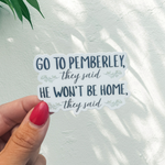 A hand with red nail polish holds a die-cut sticker that reads 'GO TO PEMBERLEY, they said HE WON'T BE HOME, they said' against a white textured wall with leaf shadows.